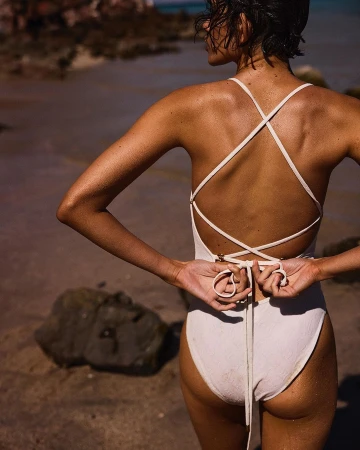 Woman wearing white one-piece swimsuit with criss-cross back straps standing on a sandy beach