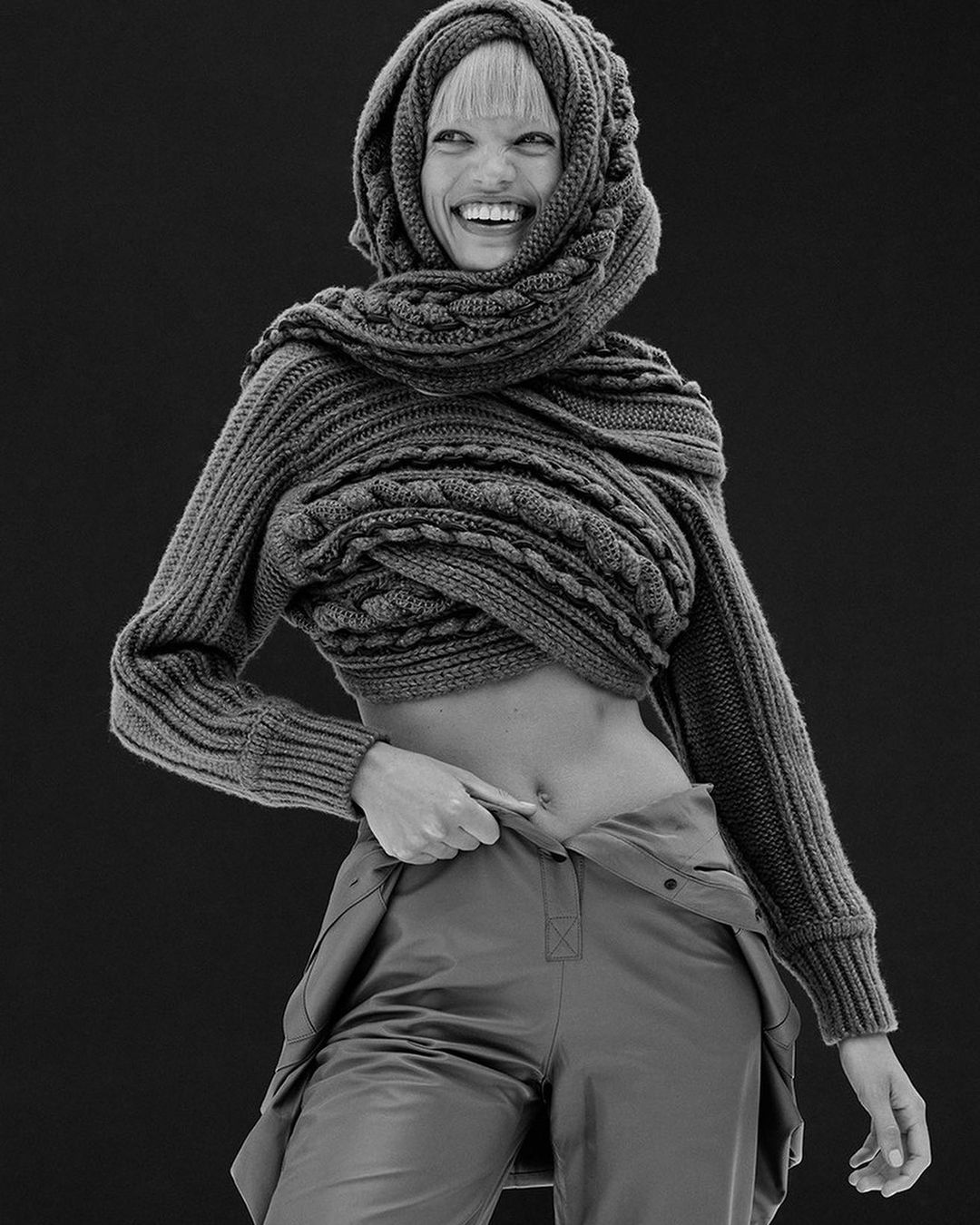 Monochrome portrait of a smiling woman with a scarf and draped knitwear, emphasizing texture and unique fashion against a dark backdrop.