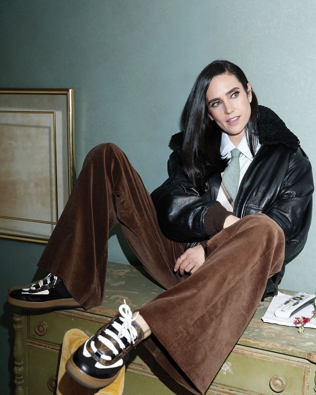 Jennifer Connelly poses indoors wearing a black bomber jacket, wide-leg brown corduroy pants, and platform sneakers, leaning on a painted dresser.
