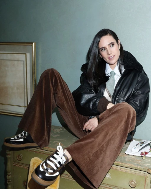 Jennifer Connelly poses indoors wearing a black bomber jacket, wide-leg brown corduroy pants, and platform sneakers, leaning on a painted dresser.