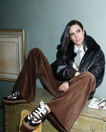 Jennifer Connelly poses indoors wearing a black bomber jacket, wide-leg brown corduroy pants, and platform sneakers, leaning on a painted dresser.