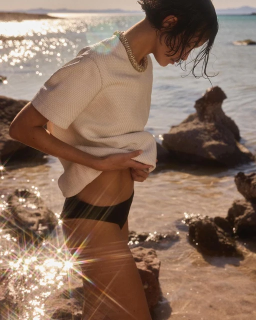 Brunette woman in a cream top and dark bikini bottoms stands near the water's edge with rocks in the background, sparkling in the sunlight.