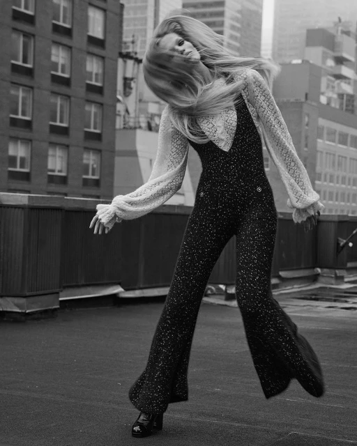 Fashion model in bell-bottom jumpsuit with sheer blouse posing on rooftop with city buildings in the background, black and white photo.