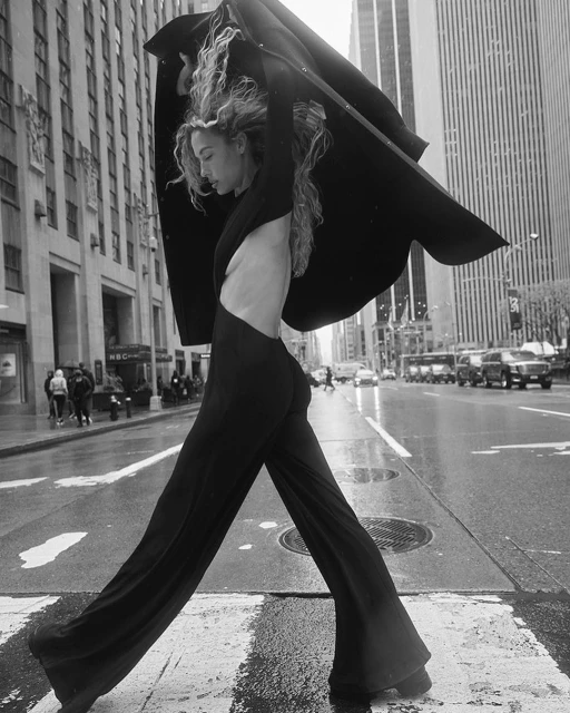 Blonde model in black clothing strides across a wet New York City crosswalk, with tall buildings in the background, fashion editorial.