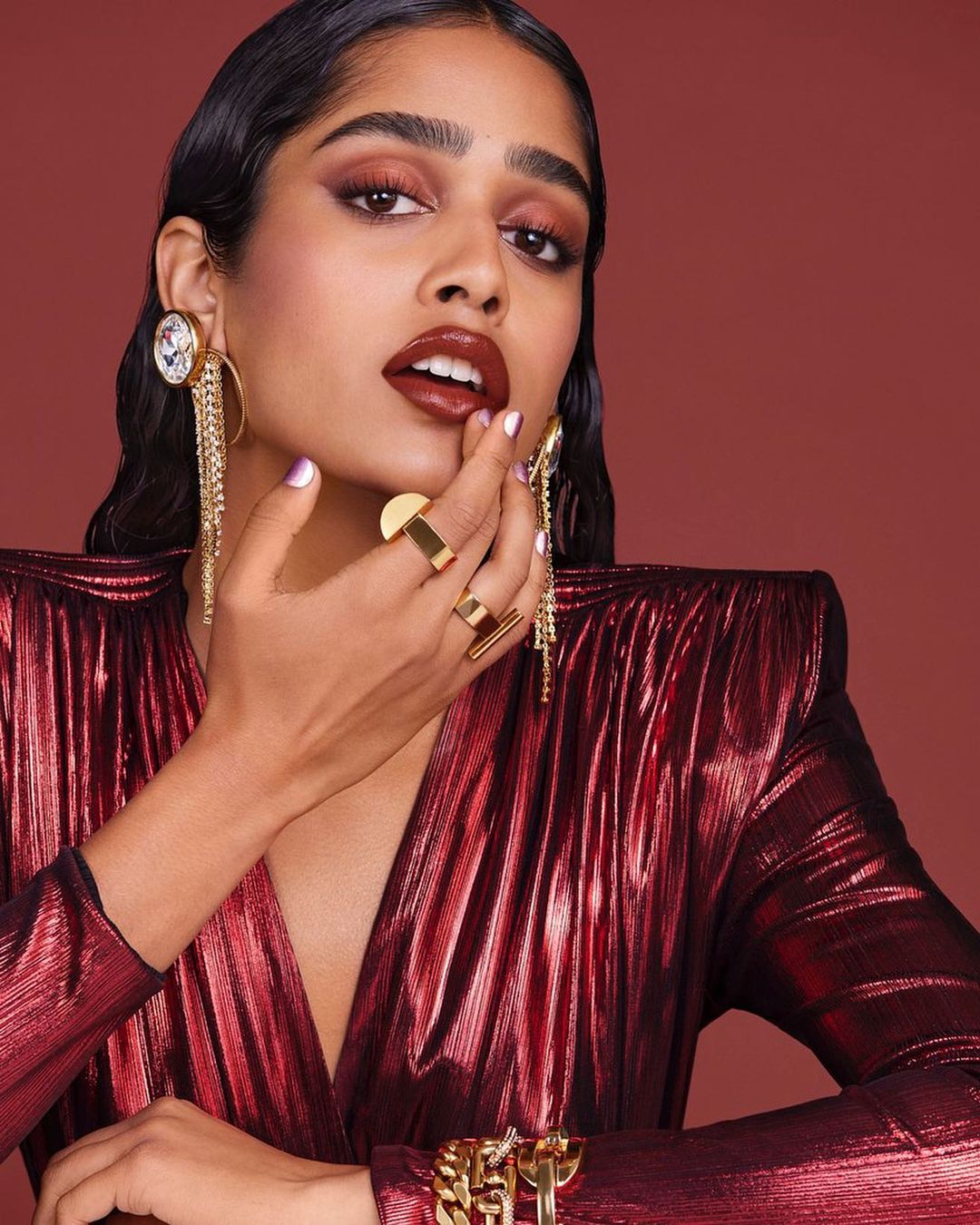 Portrait of a woman with dark hair and brown eye shadow, wearing jewelry and a sparkling red dress against a red background.