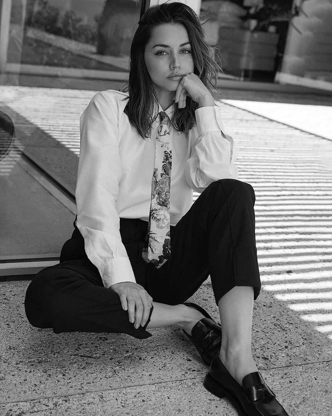 Black and white portrait of Ana de Armas seated on concrete in a white shirt and dark pants with loafers, facing the camera.