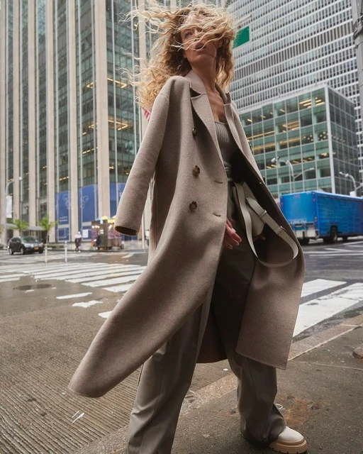 A woman in a beige coat confidently strides across a city crosswalk, with skyscrapers towering in the background and windblown blonde hair.