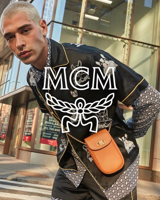 Fashionable man in printed shirt and crossbody bag poses in urban setting, showcasing modern style with a blend of casual and designer elements.