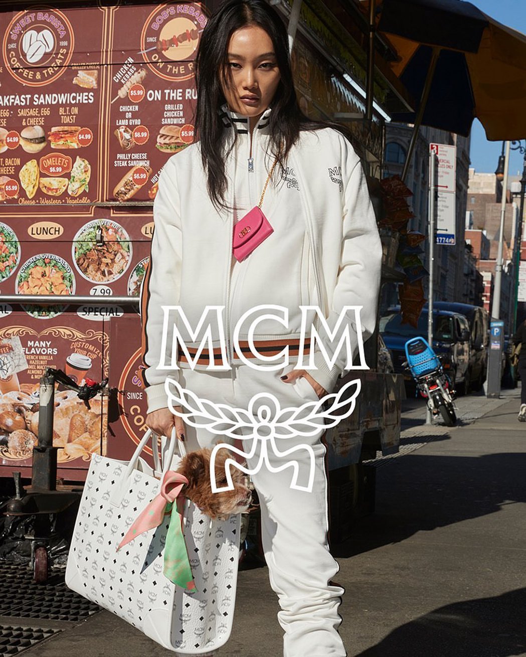 Asian female model poses outdoors in a white MCM tracksuit with a small dog peering out of a tote bag next to a food cart.