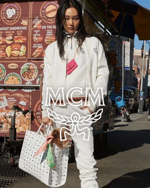 Asian female model poses outdoors in a white MCM tracksuit with a small dog peering out of a tote bag next to a food cart.