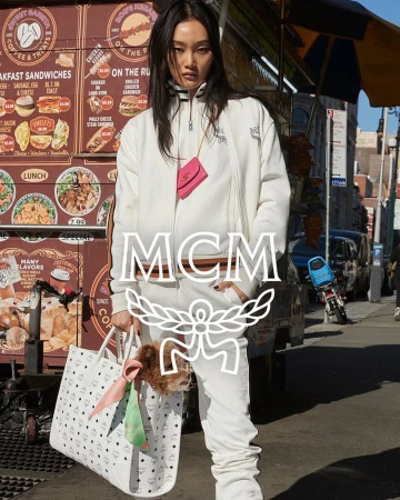 Asian female model poses outdoors in a white MCM tracksuit with a small dog peering out of a tote bag next to a food cart.