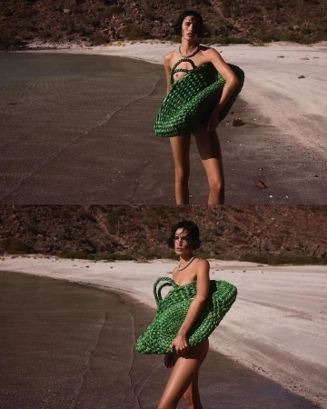 Fashion shot of a woman in swimwear on a beach holding a green woven bag. The background features an ocean and a mountain.