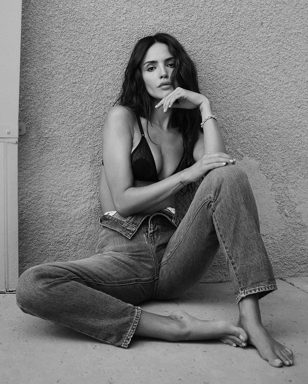 Portrait of a woman with dark hair sitting on a concrete floor wearing denim jeans and a black bra, in a black and white photo.