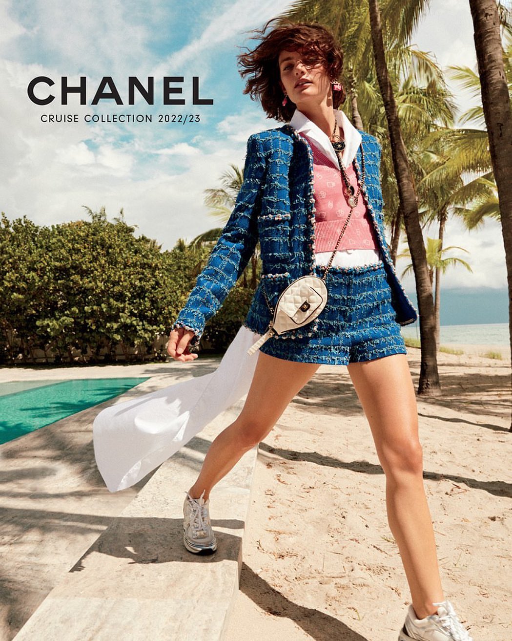 A woman models a Chanel Cruise Collection outfit on a sandy path with palm trees and a swimming pool in the background on a sunny day.