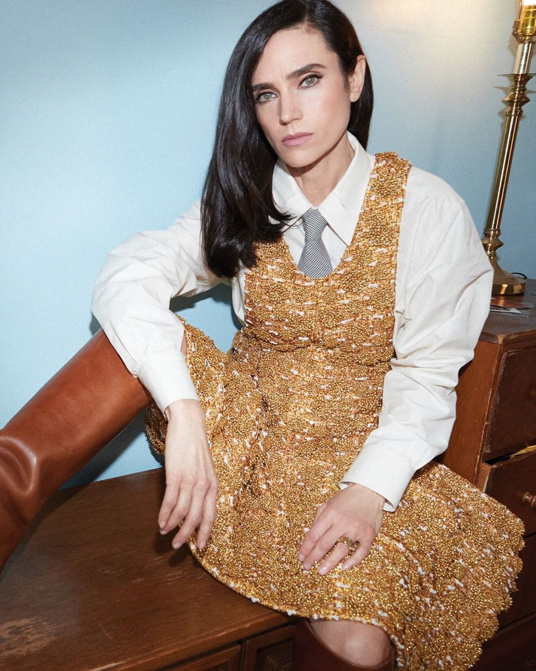 Jennifer Connelly poses in a gold textured dress with a white shirt and tie, seated indoors in a fashion style portrait.
