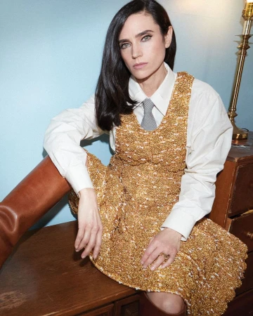 Jennifer Connelly poses in a gold textured dress with a white shirt and tie, seated indoors in a fashion style portrait.