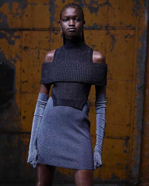 A Black female model showcases a modern, shimmering blue dress with matching long gloves in a studio with a textured background.