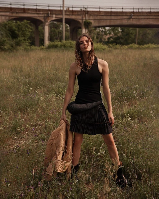 A young woman in a black dress stands in a grassy field, holding a tan jacket with an overpass in the background.