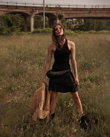 A young woman in a black dress stands in a grassy field, holding a tan jacket with an overpass in the background.