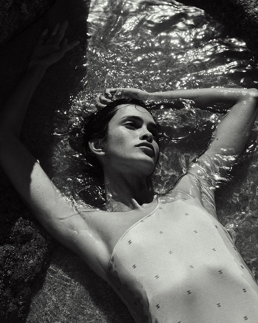 Woman in a patterned swimsuit lies in shallow water near dark rocks, looking up and away, captured in a monochrome outdoor shot.