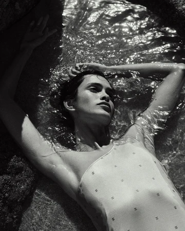 Woman in a patterned swimsuit lies in shallow water near dark rocks, looking up and away, captured in a monochrome outdoor shot.