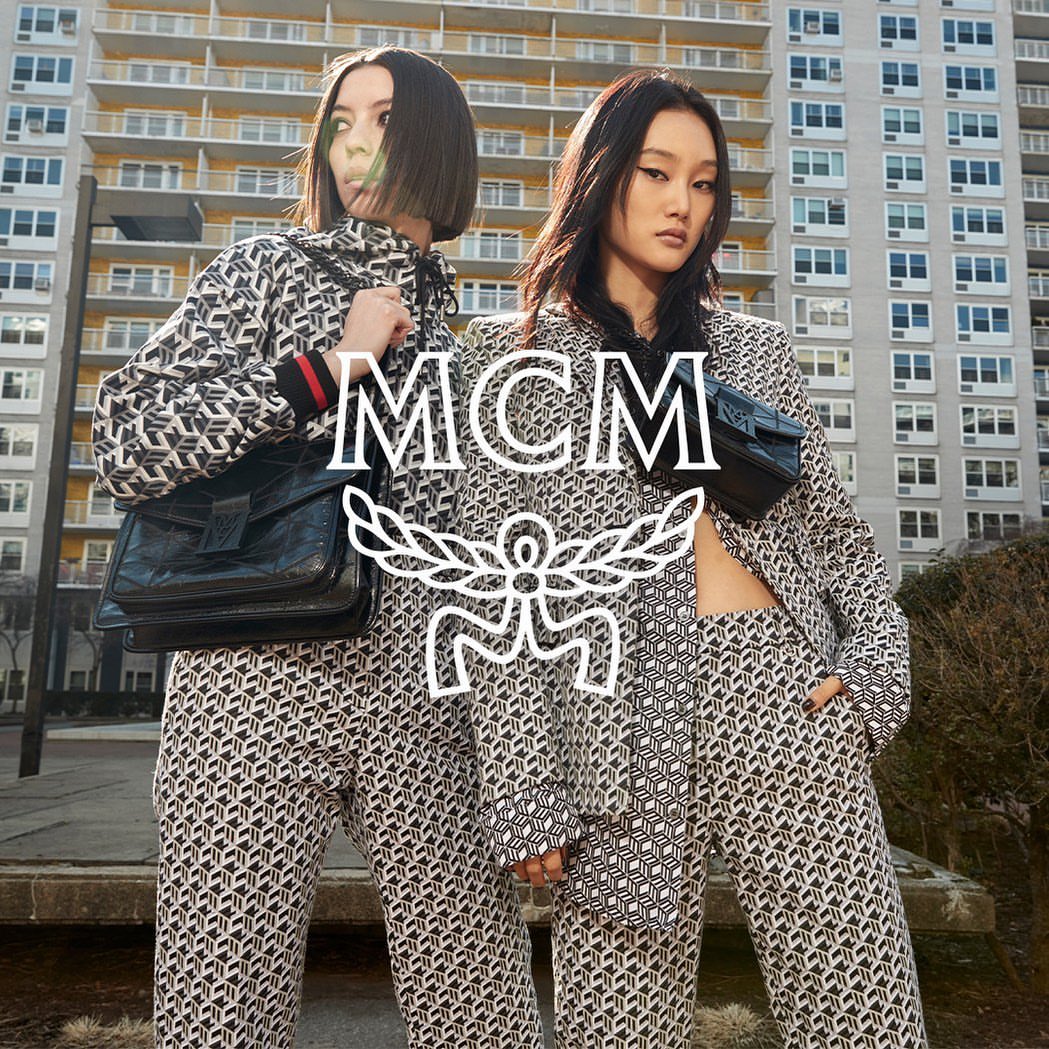 Two Asian women in patterned suits stand in front of a building, holding MCM brand purses. A large MCM logo is superimposed.