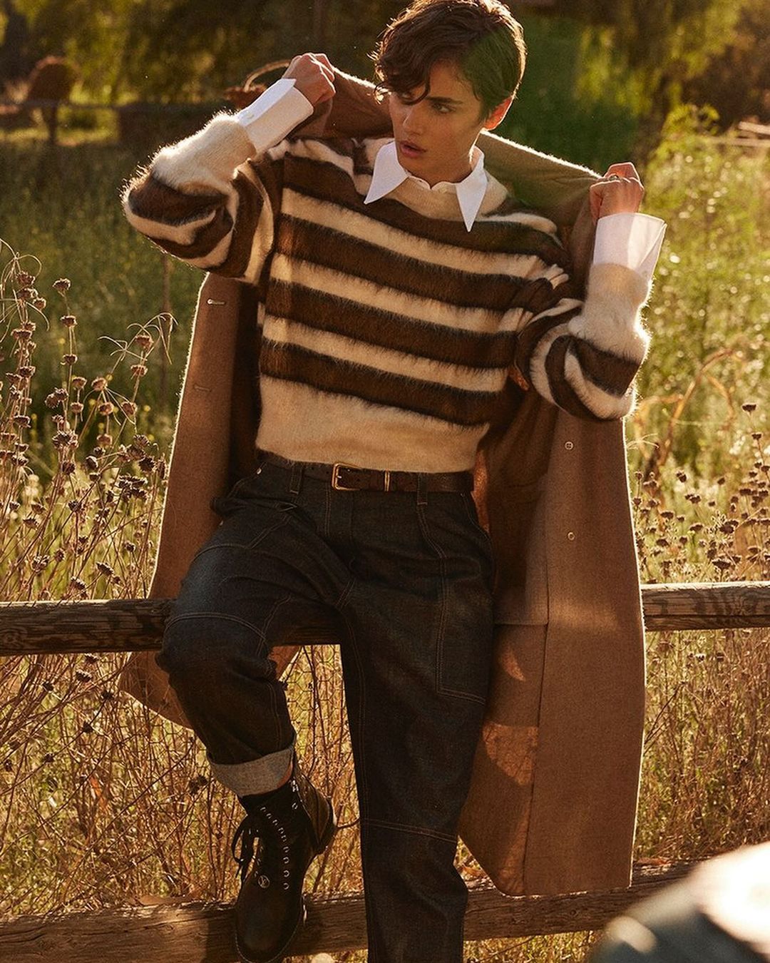 Young adult in striped sweater and dark jeans leans against wooden fence outdoors in natural sunlight with trees in background.
