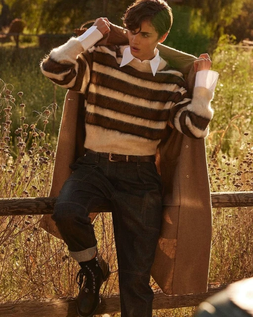 Young adult in striped sweater and dark jeans leans against wooden fence outdoors in natural sunlight with trees in background.