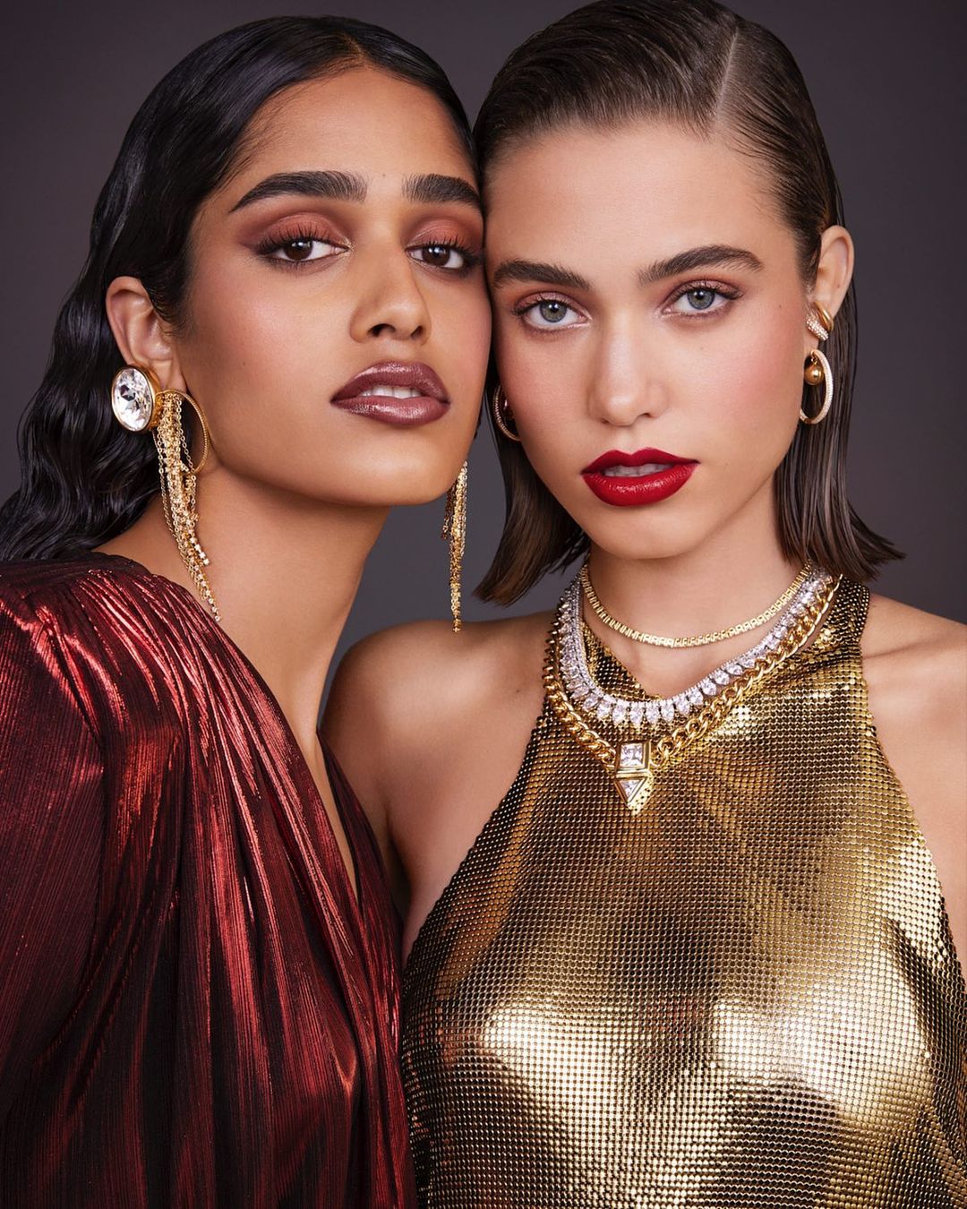 Two glamorous women pose in a studio, adorned in golden and red metallic dresses and sparkling jewelry, embodying elegance and style.