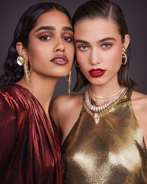 Two glamorous women pose in a studio, adorned in golden and red metallic dresses and sparkling jewelry, embodying elegance and style.