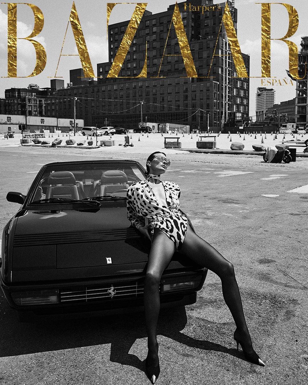 Fashion editorial with a woman in a leopard print bodysuit posing on a black Ferrari convertible in an urban setting with buildings.