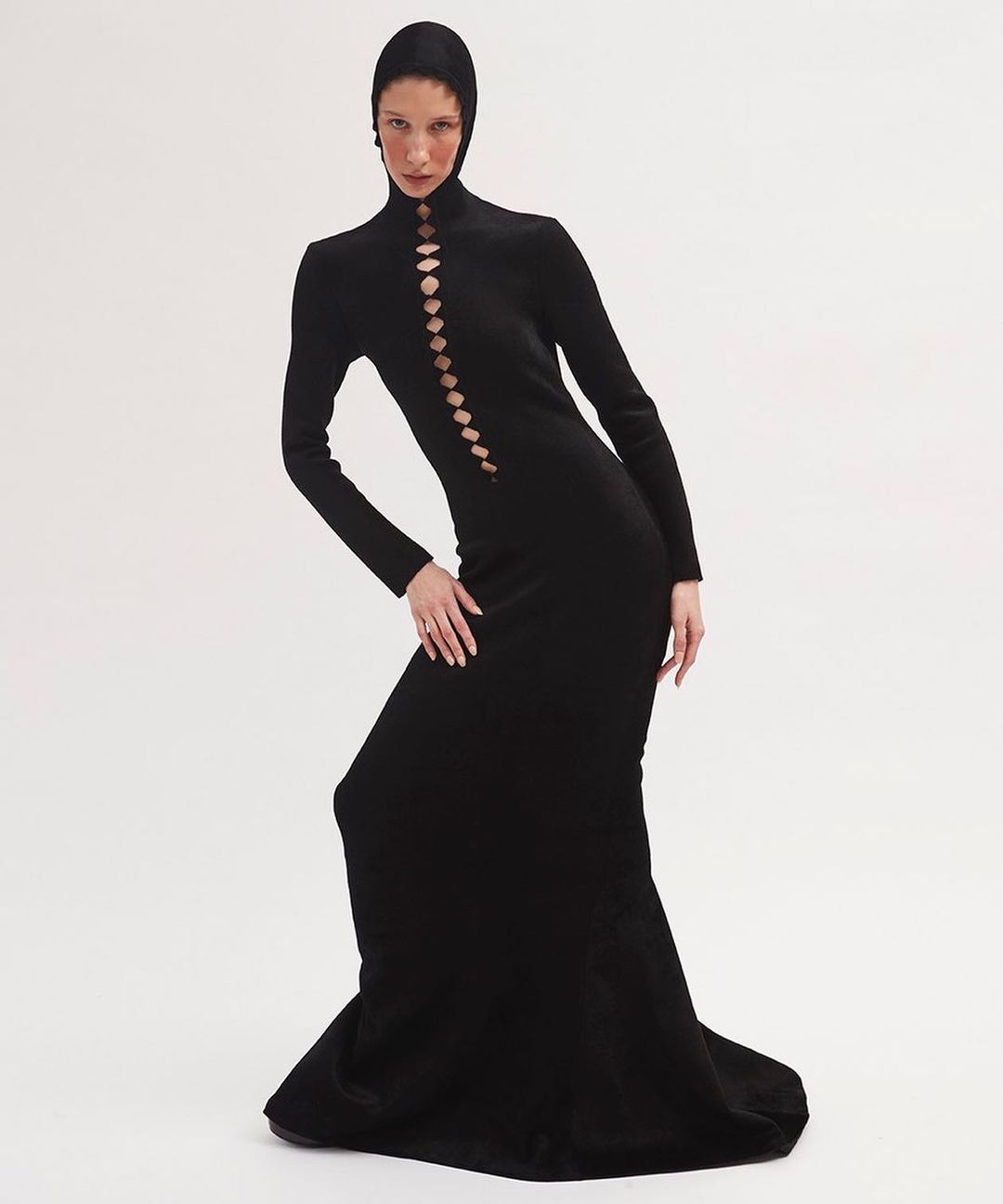 An adult female fashion model poses in a black dress with cut-out details against a plain white backdrop in a studio fashion shot.