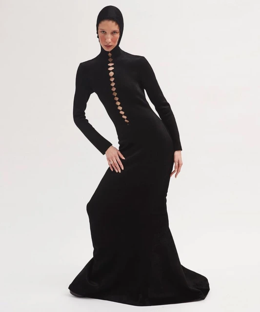 An adult female fashion model poses in a black dress with cut-out details against a plain white backdrop in a studio fashion shot.