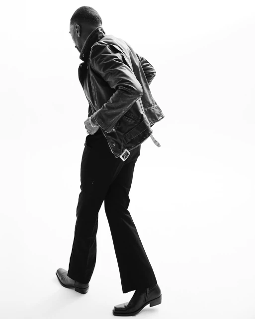 Black and white portrait of a man in a leather jacket walking against a white background, showcasing fashion and monochrome aesthetics.