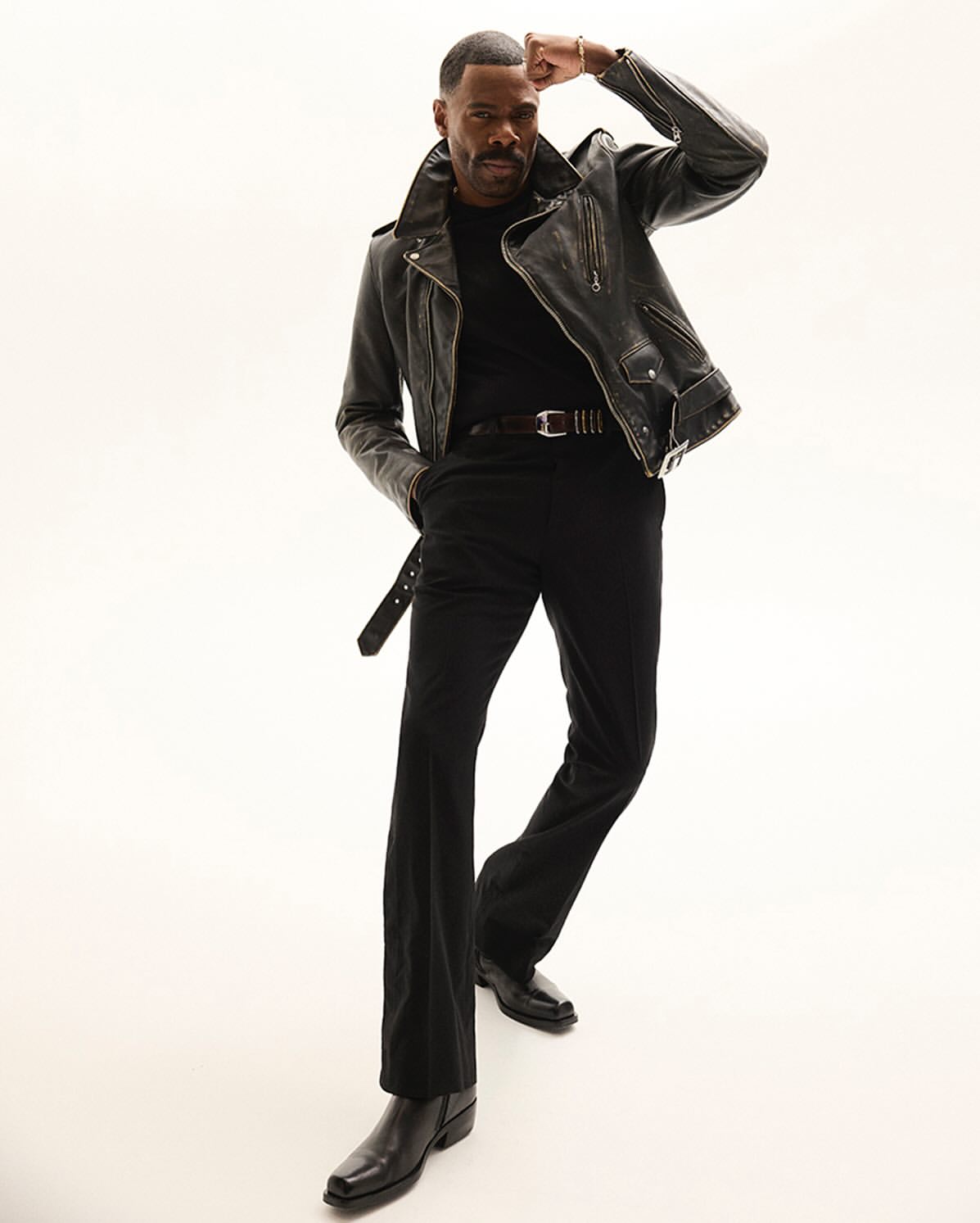 Full-length portrait of African-American man in leather jacket, black pants and boots, posing against white background in a studio.