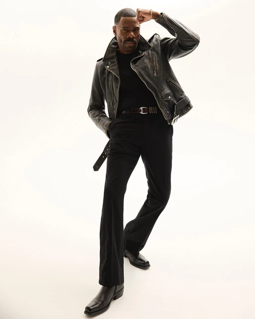 Full-length portrait of African-American man in leather jacket, black pants and boots, posing against white background in a studio.