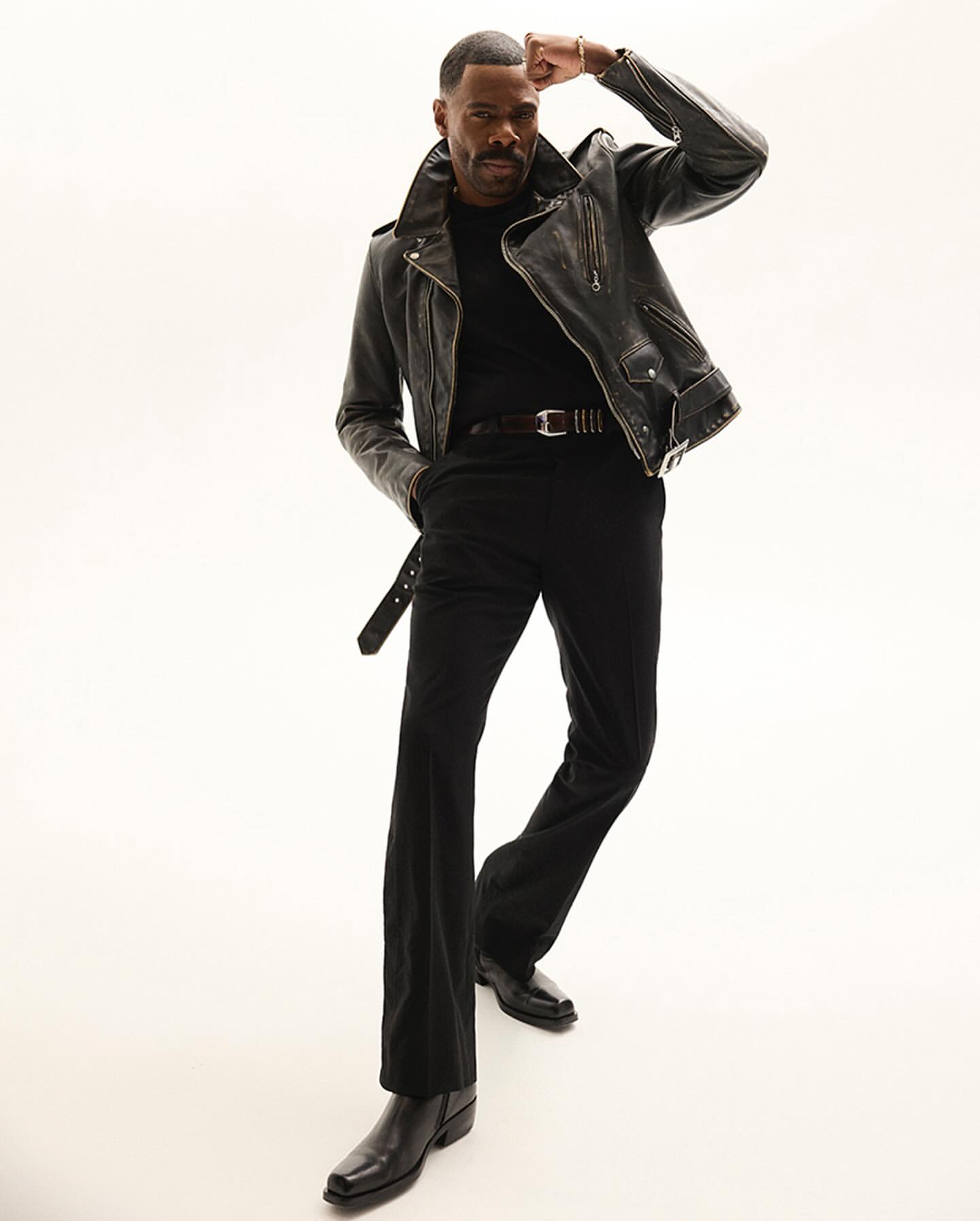 Portrait of a Black man in a leather jacket and black outfit, posing confidently in a full-body shot against a plain white backdrop.