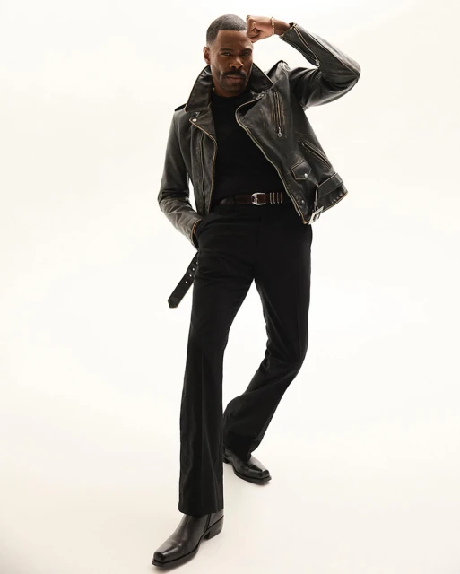 Portrait of a Black man in a leather jacket and black outfit, posing confidently in a full-body shot against a plain white backdrop.