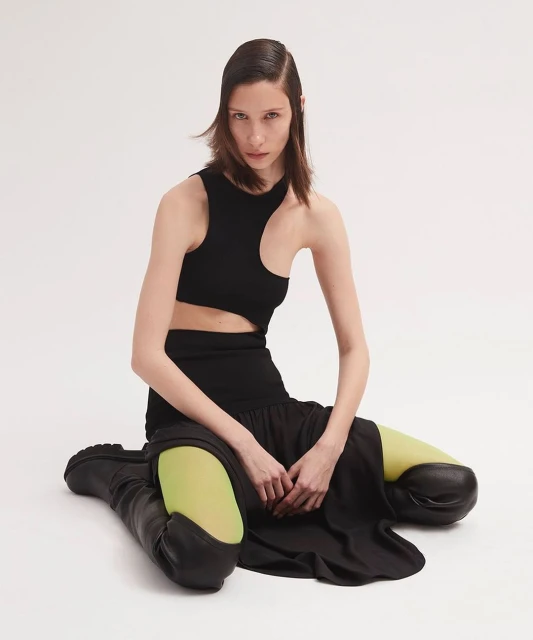 A woman with dark hair poses in a black cut-out top, black skirt, black boots, and chartreuse tights against a bright white backdrop.