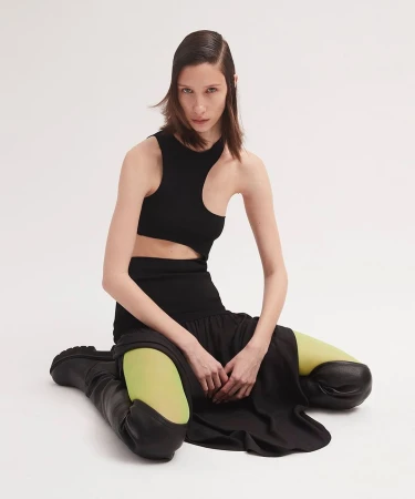 A woman with dark hair poses in a black cut-out top, black skirt, black boots, and chartreuse tights against a bright white backdrop.