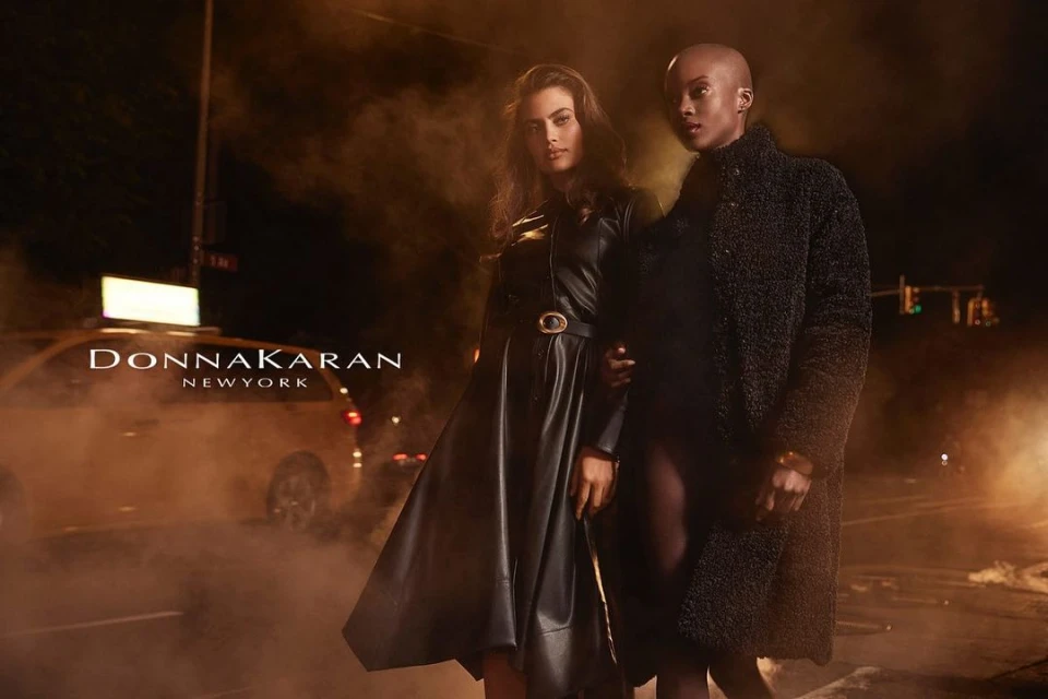Two women pose in Donna Karan New York ad wearing black coats on a city street at night, taxi, street lights, and foggy haze.