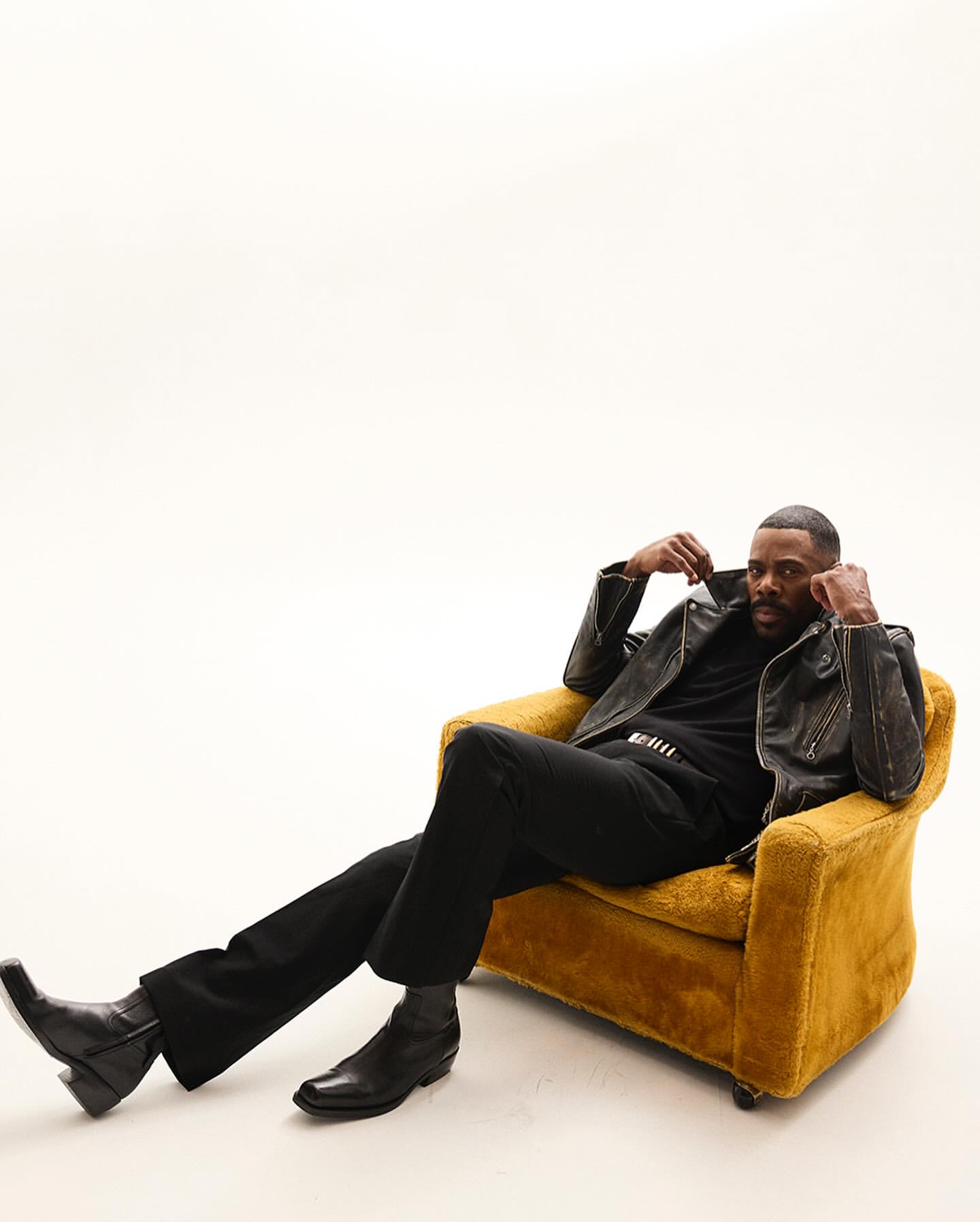 An adult Black male is seated in a yellow arm chair, wearing a black leather jacket, black pants, and black boots, against a white backdrop.