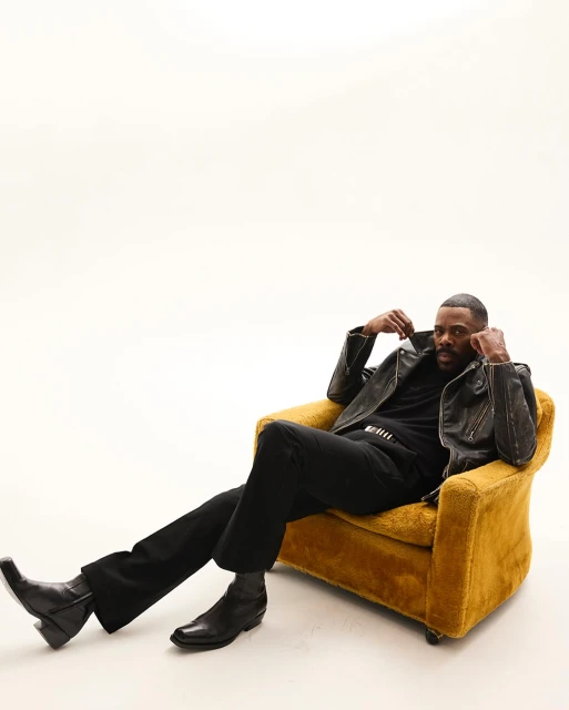 An adult Black male is seated in a yellow arm chair, wearing a black leather jacket, black pants, and black boots, against a white backdrop.