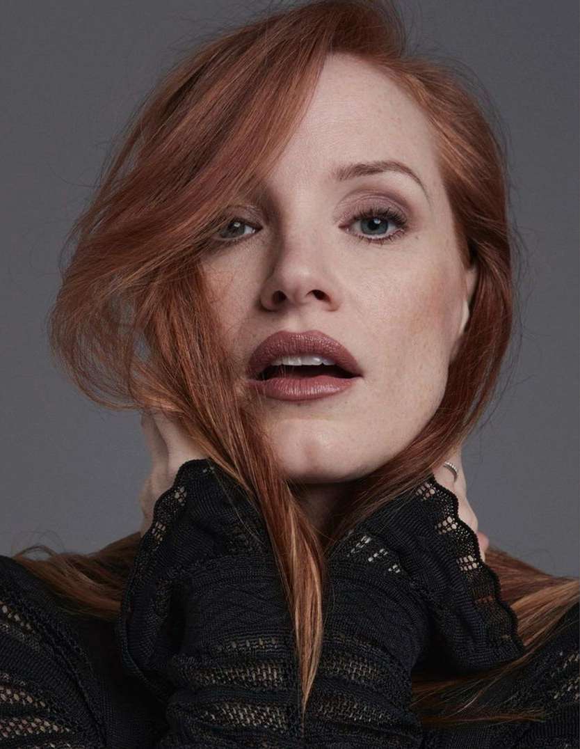 Portrait of a red-haired woman in a black textured top. She is holding her hands at her neck in an indoor studio setting.