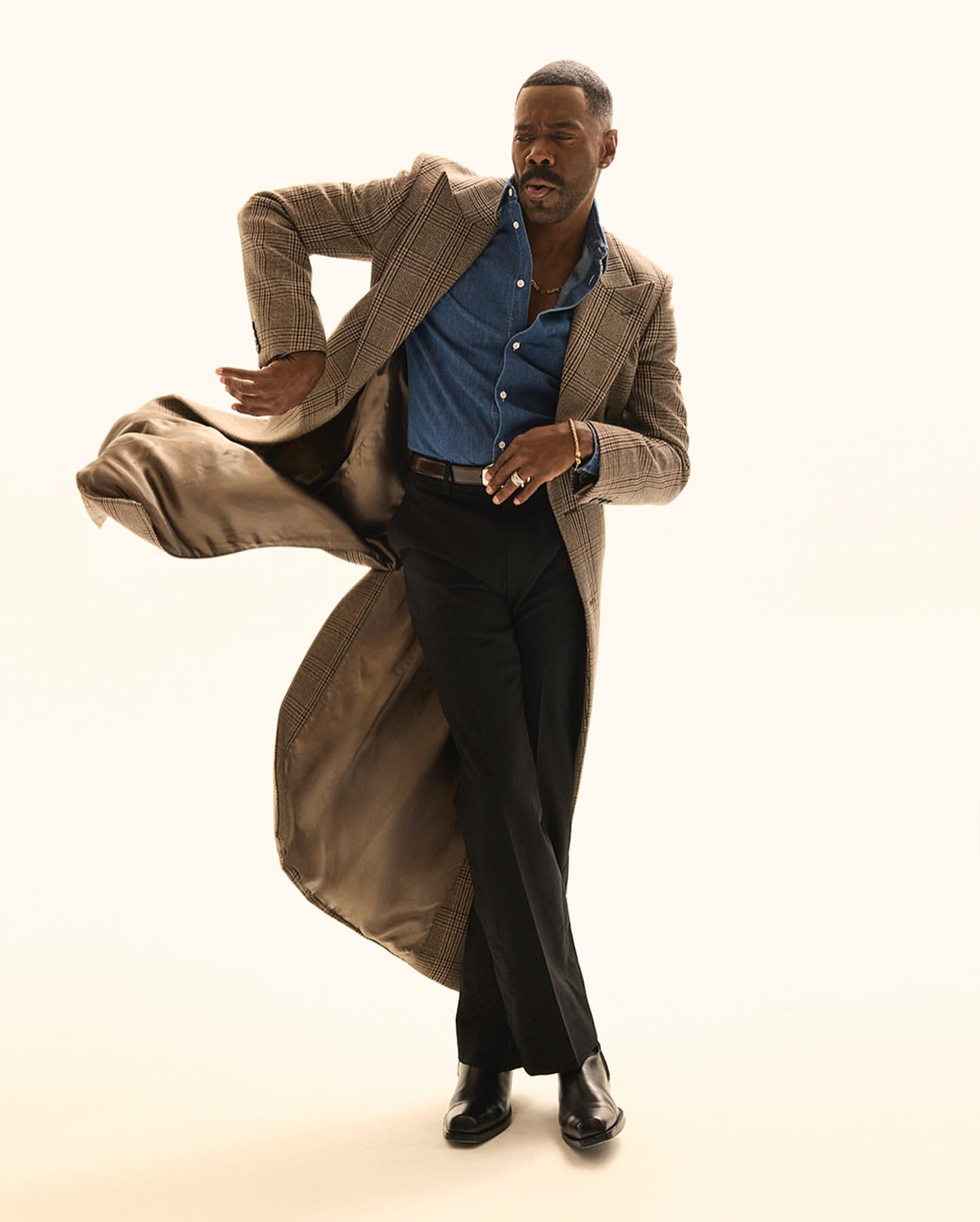 Stylish adult male in a coat, jeans shirt, and dress pants strikes a dynamic pose against a neutral background, showcasing fashion and style.