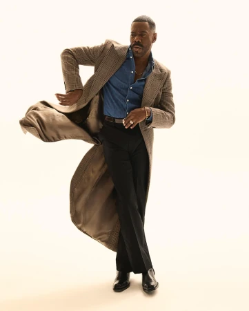 Stylish adult male in a coat, jeans shirt, and dress pants strikes a dynamic pose against a neutral background, showcasing fashion and style.