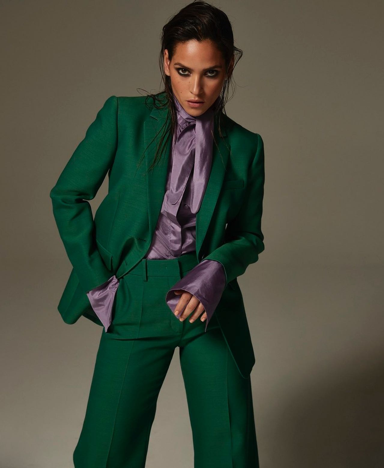 Brunette woman in a green suit and lavender silk blouse poses against a grey backdrop in a fashion forward studio portrait.