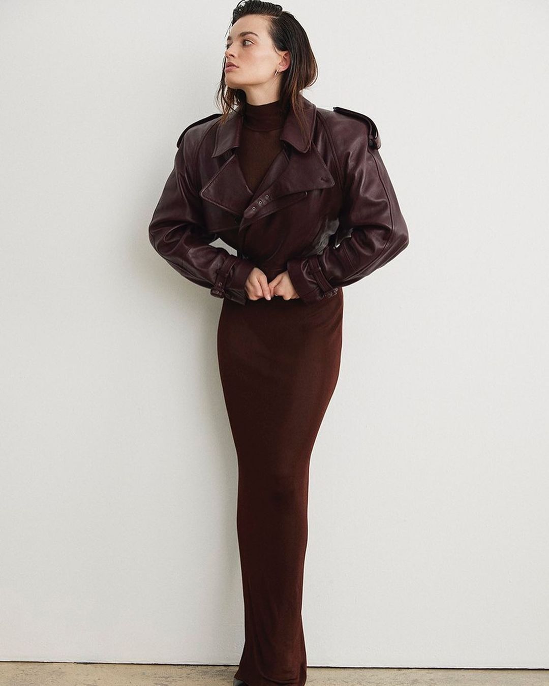 A woman in a long brown dress and brown leather jacket poses against a white wall in a studio shot with soft, diffused lighting.