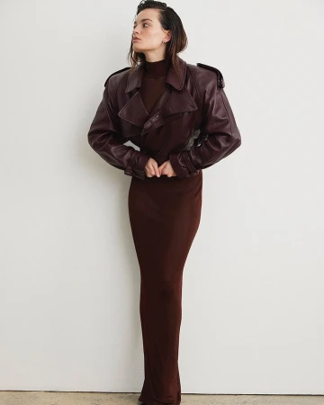 A woman in a long brown dress and brown leather jacket poses against a white wall in a studio shot with soft, diffused lighting.