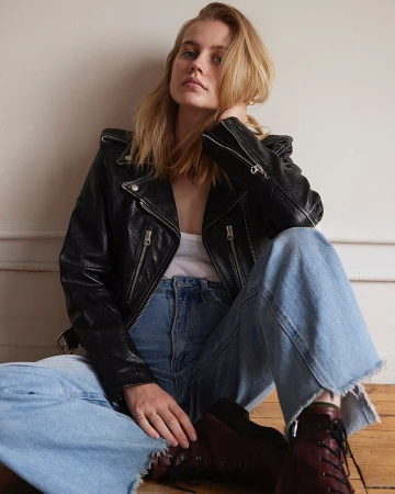 Portrait of a blonde woman wearing a black leather jacket and blue jeans seated against a wall
